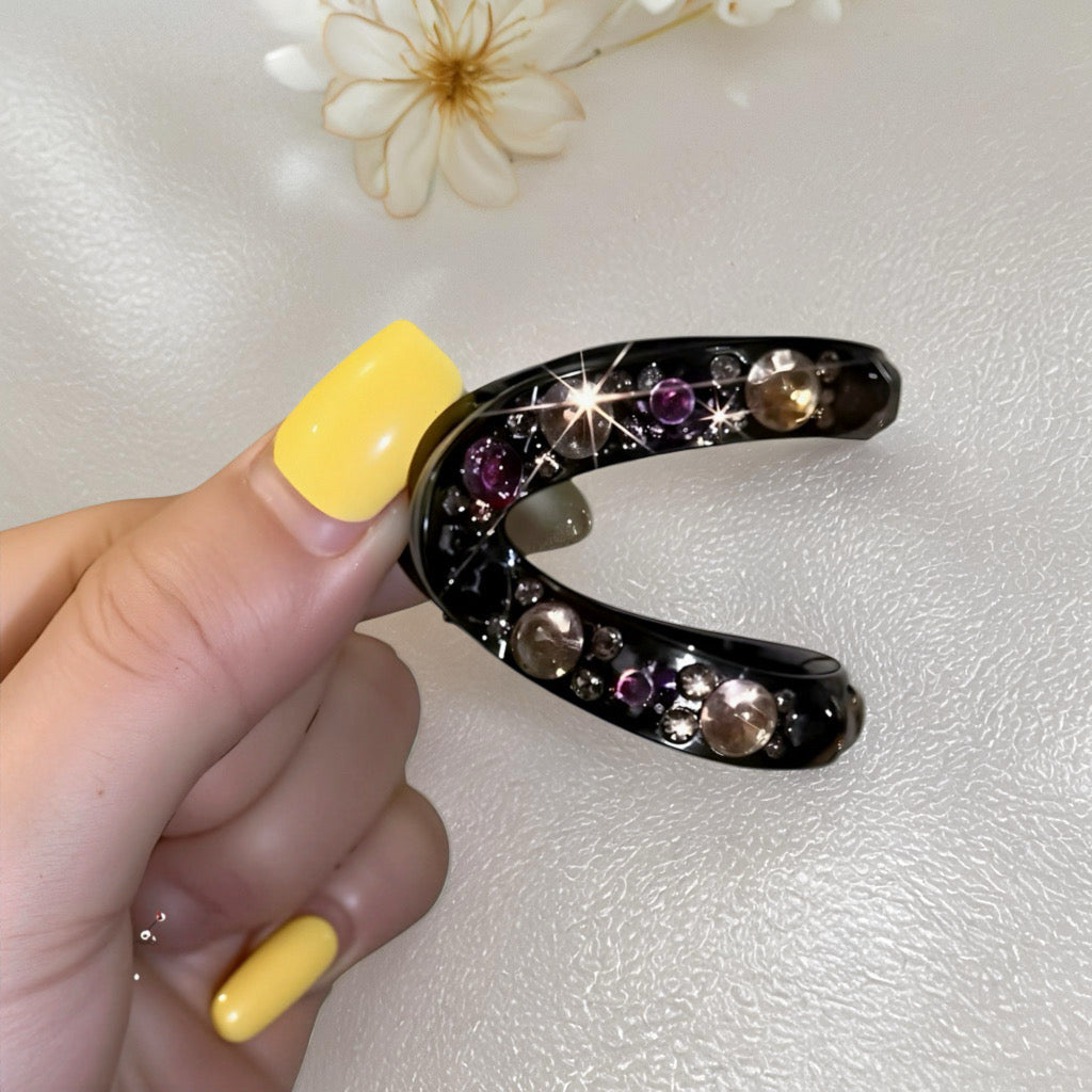 Hand holding black crescent crystal hair claw with sparkling red and champagne gems, nail art visible.