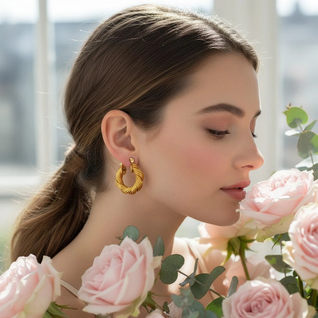 PaulaB Jewellery model wearing Twisted 18k gold-plated copper hoop earrings among soft pink roses, feminine and Parisian inspired luxury earrings for women in Australia, NZ & UK.